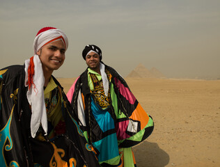 Fototapeta premium Two cheerful young Egyptian folk dancers in colorful tanoura costumes and turbans smiling before the Giza pyramids ideal for cultural festival and