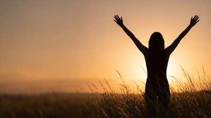 Fototapeta premium Silhouette of a woman with arms raised in tall grass against a warm orange sunset, faceless backlit figure of freedom with wide left copy space