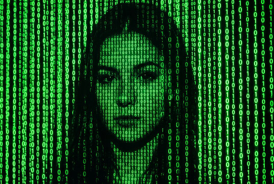 Digital illustration of a human face hidden behind a stream of green binary code data. A digitized female face embedded in a dense stream of green binary code on a black background