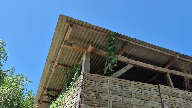 Low angle panning shot of a traditional hut corner with woven bamboo walls and corrugated zinc roof against a clear blue sky