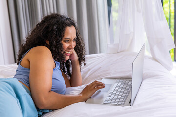 African American woman reclining on bed, propping on left elbow, smiling while using silver laptop © wavebreak3