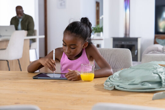 Father and child sitting at wooden table, using stylus on tablet, orange juice, backpack, laptop