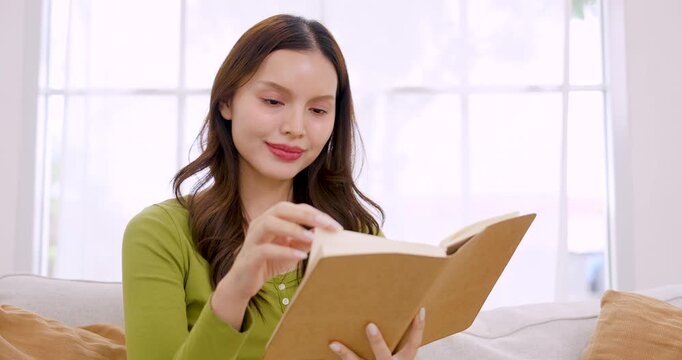 Young Asian woman reads book on cozy couch, traces page with finger, smiles gently, then keeps reading, creating calm home lifestyle mood for weekend hobby, self care, indoor relaxation time at home
