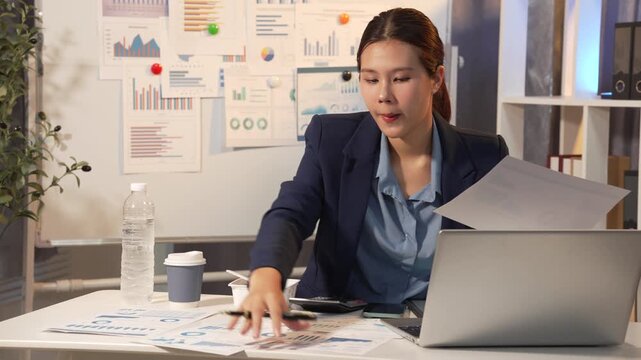 Businesswoman reviewing charts and documents at a desk in a modern office with laptop, coffee, and water bottle