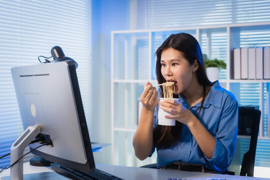 Young woman eating instant noodles at her desk while working on a computer in a modern office at night, showing overtime, busy schedule, and quick meal during work