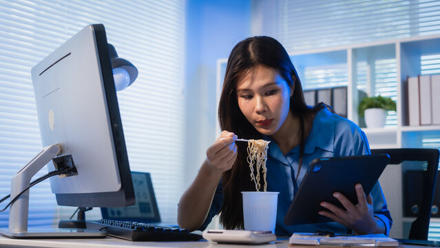 Young woman eating instant noodles at her desk while working on a computer in a modern office at night, showing overtime, busy schedule, and quick meal during work