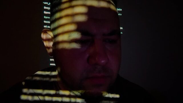Computer hacker working in dark room with green programming code projected on his face, cyber security concept