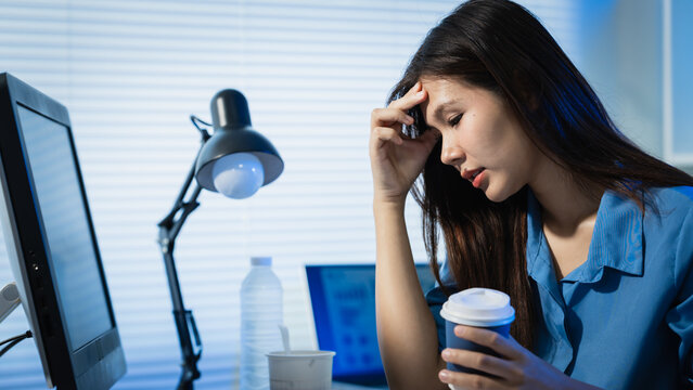 Tired young woman sitting at office desk with coffee, holding her head beside a computer, showing stress, fatigue, burnout and overwork in a late work setting.