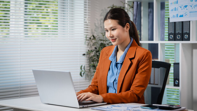 Smiling young businesswoman working on a laptop at her office desk, with reports, shelves and window blinds in the background, showing a calm and productive workday.
