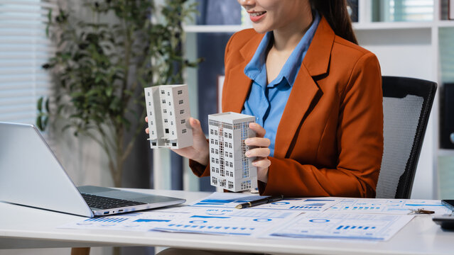 Office desk with a small building model, calculator, charts and tablet, while a businesswoman reviews a property sample, showing real estate planning, finance and investment work.