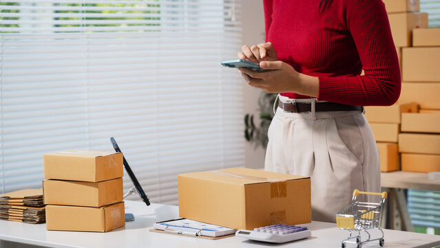 Woman in a home office packing parcel boxes and using a smartphone at a work table, showing small business shipping, online orders, and e-commerce fulfillment.