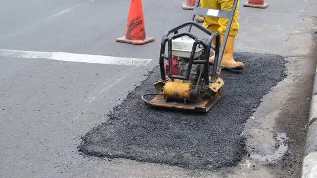 a road worker using a plate compactor to flatten fresh asphalt patch during street repair and maintenance.