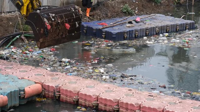 an excavator bucket draining water while removing plastic trash and debris from a polluted urban canal.
