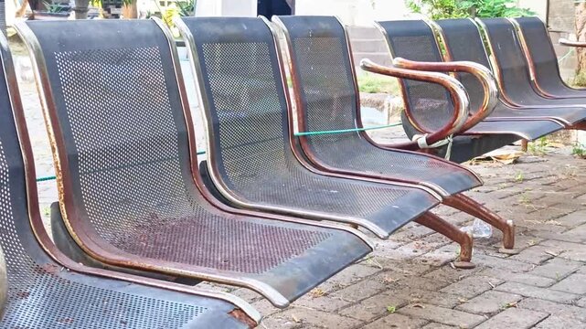 Row of empty perforated metal waiting chairs outdoors on a paved public area.