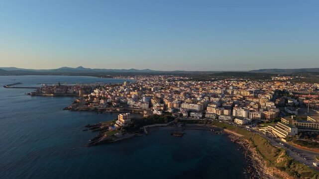 Alghero coastal skyline glows at golden hour as a drone flies along the rocky shoreline and dense city.