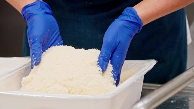 Professional food service worker wearing blue gloves mixing a large quantity of grated cheese in white industrial kitchen containers