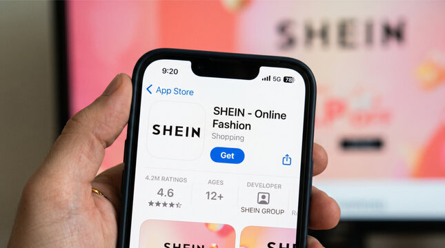 SHEIN online fashion shopping app interface on an iPhone screen held by a hand. The App Store page displays the logo, ratings, and download button with a blurred SHEIN website in the background.
