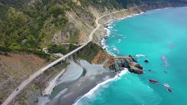 Aerial drone view of the iconic Bixby Creek Bridge spanning a canyon along Highway 1 with turquoise ocean water below.