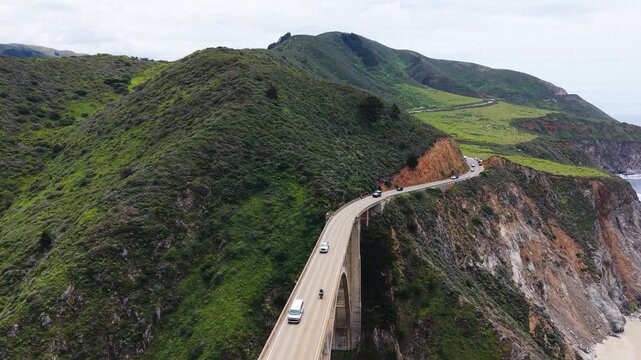 Aerial drone view of cars crossing the historic Bixby Creek Bridge surrounded by green hills in Big Sur.