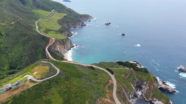 Aerial drone view of Highway 1 winding along steep green cliffs above turquoise water and offshore sea stacks in Big Sur.