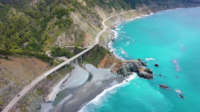 Aerial drone view of the historic Bixby Creek Bridge spanning a canyon above a sandbar and turquoise ocean water.