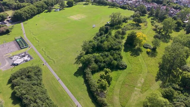 Drone orbit of crescent trees at Chad park, Chadwell Heath, showing grass, cricket field, paths and woodland edge on a bright summer day in suburban East London, UK.