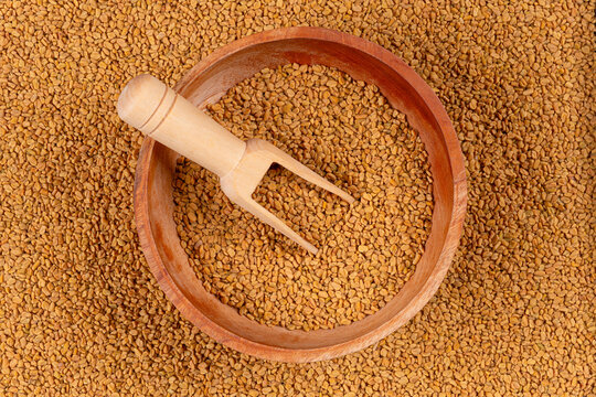 Top view of a wooden bowl filled with fenugreek seeds and a wooden scoop on a fenugreek seed background