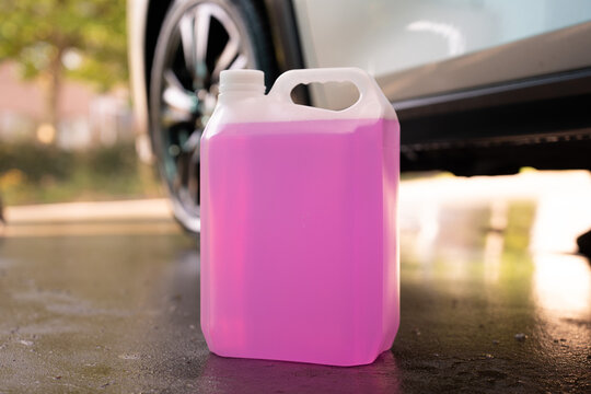 Plastic canister with pink liquid stands on wet ground near car wheel close up. Windshield washer fluid, antifreeze coolant or car shampoo bottle, auto maintenance and detailing concept.
