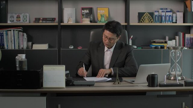 A businessman in a suit sitting at an executive desk writing on documents with a laptop open beside him and bookshelves in the background at night.