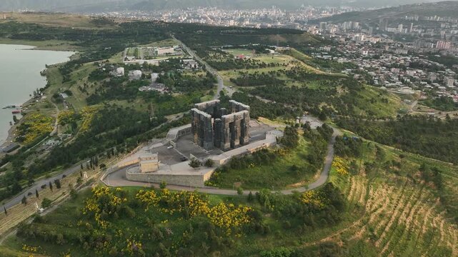 Drone shot capturing the historical Chronicle of Georgia monument complex during a scenic evening sunset. The huge stone pillars are set on a green hill next to the calm Tbilisi Sea reservoir