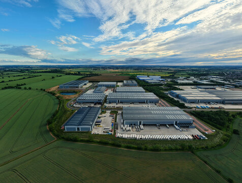 Aerial view of a logistics park with warehouse buildings, loading docks, parked semi-truck trailers, and surrounding green fields in Biggleswade, England, United Kingdom.