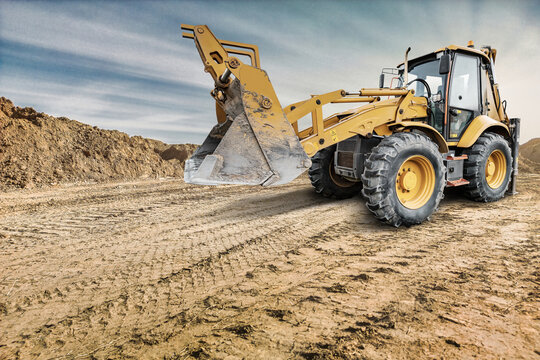 Universal excavator loader digs into the ground at a construction location with dirt piles nearby while preparing for other tasks on site during daylight hours