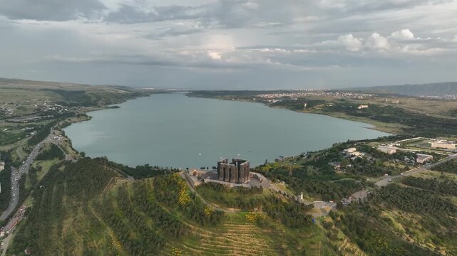 Drone shot capturing the historical Chronicle of Georgia monument complex during a scenic evening sunset. The huge stone pillars are set on a green hill next to the calm Tbilisi Sea reservoir