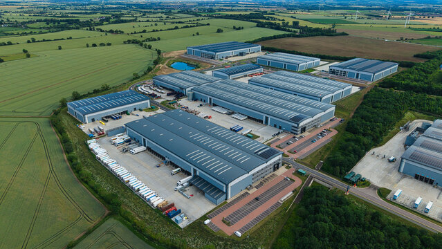 Aerial view of a large logistics park with warehouses, loading docks, and trucks parked in rows, surrounded by green fields in Biggleswade, England, United Kingdom.