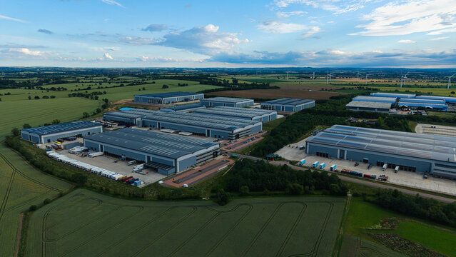 Aerial view of a large industrial logistics park featuring multiple warehouses, loading docks, and parked trucks surrounded by green fields in Biggleswade, England, United Kingdom.