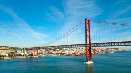 Fototapeta premium Red suspension bridge connecting both sides of the river estuary, showcasing urban landscape and a large container port activity under a bright blue sky