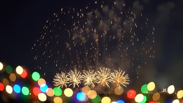 Fireworks explode in the night sky above a festive display of colorful blurred string lights.