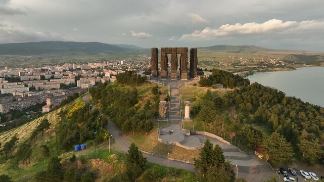 Drone shot capturing the historical Chronicle of Georgia monument complex during a scenic evening sunset. The huge stone pillars are set on a green hill next to the calm Tbilisi Sea reservoir