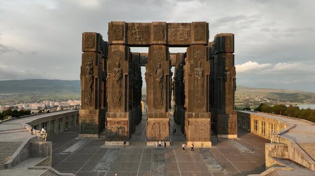 Drone shot capturing the historical Chronicle of Georgia monument complex during a scenic evening sunset. The huge stone pillars are set on a green hill next to the calm Tbilisi Sea reservoir