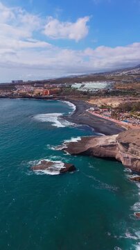 Aerial view of black sand beach, rocky coastline, and resort buildings on a sunny day
