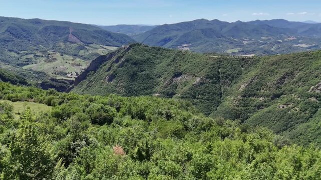 Ampio paesaggio montano ricoperto di vegetazione verde, con valli e creste che si estendono in lontananza sotto un cielo azzurro limpido. Scena naturale vasta e luminosa.
