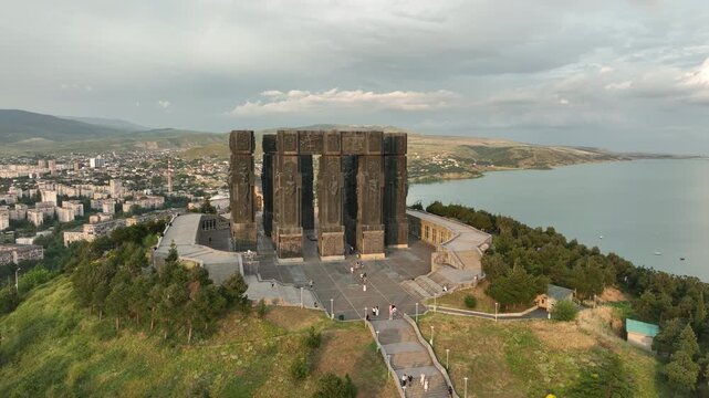 Drone shot capturing the historical Chronicle of Georgia monument complex during a scenic evening sunset. The huge stone pillars are set on a green hill next to the calm Tbilisi Sea reservoir