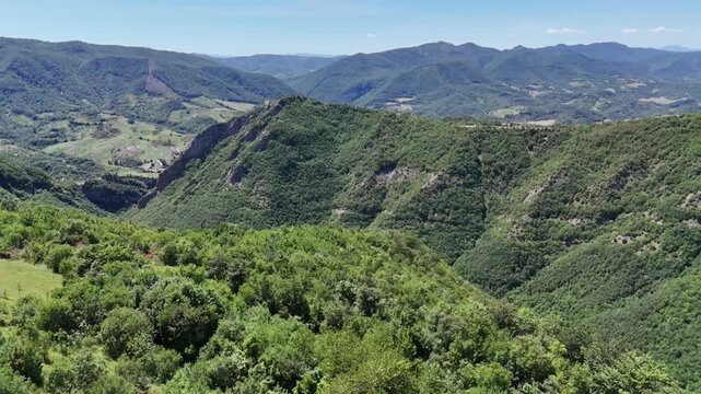 Ampio paesaggio montano ricoperto di vegetazione verde, con valli e creste che si estendono in lontananza sotto un cielo azzurro limpido. Scena naturale vasta e luminosa.