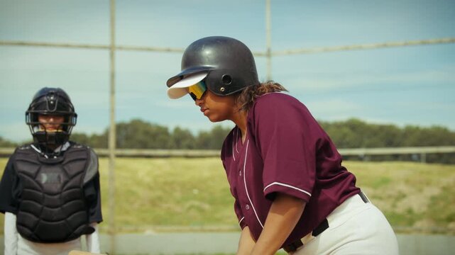 A focused female batter in a maroon jersey and white pants takes her stance at home plate, ready to swing. The catcher is visible behind her, awaiting the pitch.