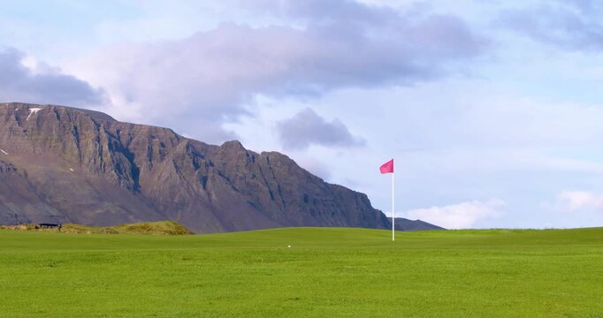 A red flag marks the hole on a pristine green golf course, set against a backdrop of a majestic, rugged mountain range under a cloudy sky.