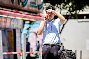 Fototapeta premium 屋外でスマートフォンで通話しながら日差しを遮る夏バテの仕事中の半袖の30代の日本人の男性の会社員