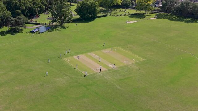 Chat Park Chadwell Heath drone flyover showing local cricket match on a sunny summer day, wide aerial view of green fields, pitch, players, and surrounding suburban treeline in London, UK.