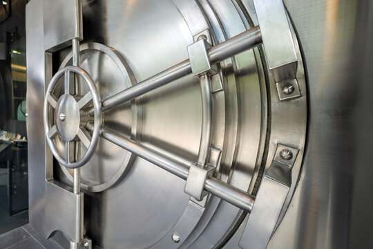 A close-up shot of a heavy-duty, circular stainless steel bank vault door showcasing its intricate mechanical locking wheel, security bolts, and solid metallic structure.