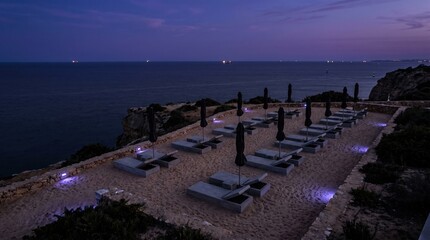 Obraz premium The scene shows a beach lounge area with many seats and umbrellas placed on the sand. Lights glow softly as the sun sets over the ocean, creating a relaxed atmosphere.