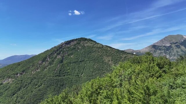 Montagna verde ricoperta da vegetazione fitta, con sentiero visibile sul versante e cime sullo sfondo sotto un cielo azzurro limpido